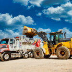 yellow earthmoving tractor dumping concrete material into proall mixer with blue clouded sky background